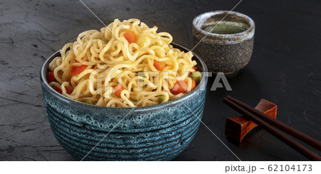 Instant noodles panorama, a bowl of soba with carrot and scallions, with a cup of sake and chopsticks, on a black background Instant noodles panorama, a bowl of soba with carrot and scallions, with a cup of sake and chopsticks, on a black background 62104173