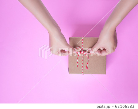 Female hands bow tie a simple craft paper gift box wrapped with colorful rope with as a present for Christmas, new year, valentine day or anniversary on pink background, top view Female hands bow tie a simple craft paper gift box wrapped with colorful rope with as a present for Christmas, new year, valentine day or anniversary on pink background, top view 62106532