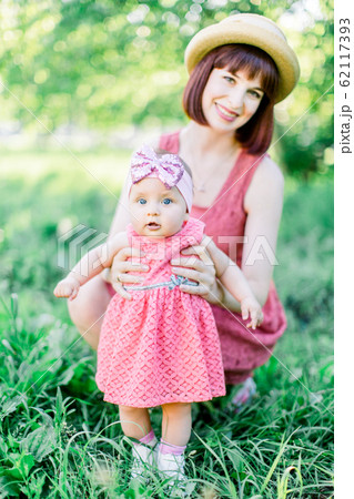 Cute funny happy baby making his first steps on a green grass, mother holding her hands supporting by learning to walk Cute funny happy baby making his first steps on a green grass, mother holding her hands supporting by learning to walk 62117393