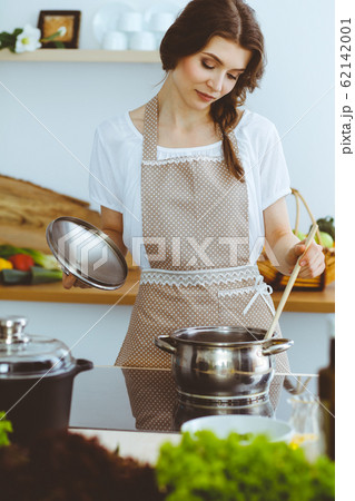 Young brunette woman cooking soup in kitchen. Housewife holding wooden spoon in her hand. Food and health concept Young brunette woman cooking soup in kitchen. Housewife holding wooden spoon in her hand. Food and health concept 62142001