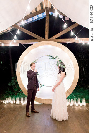Night wedding ceremony outdoors. Bride and groom swear an oath each other on wedding arch background. Beautiful just married couple. Rustic wooden restaurant with light bulbs 62144802