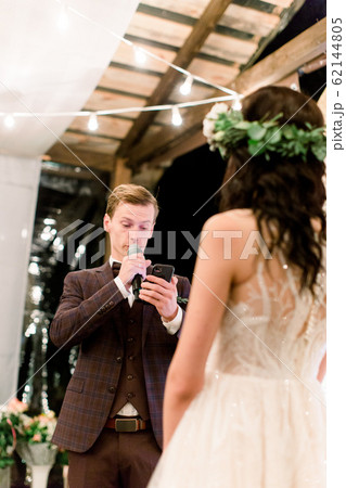 Side view of stylish groom reading his oath to the bride at a ceremony near the wedding arch, outdoors, standing in wooden tent restaurant decorated with light bulbs. Rustic wedding Side view of stylish groom reading his oath to the bride at a ceremony near the wedding arch, outdoors, standing in wooden tent restaurant decorated with light bulbs. Rustic wedding 62144805
