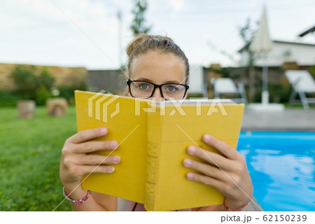Teenage girl in glasses reads a book, background swimming pool, lawn near the house. School, education, knowledge, adolescents Teenage girl in glasses reads a book, background swimming pool, lawn near the house. School, education, knowledge, adolescents 62150239