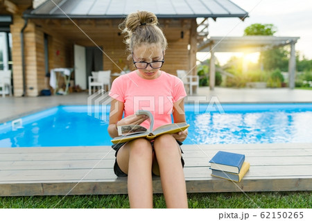 Young girl in glasses near the pool with a pile of books, reading book. Education, summer, knowledge 62150265