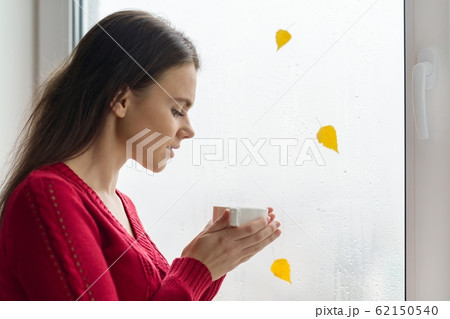 Autumn in the window, a young girl stands near the window with raindrops and a yellow leaf, drinking coffee Autumn in the window, a young girl stands near the window with raindrops and a yellow leaf, drinking coffee 62150540
