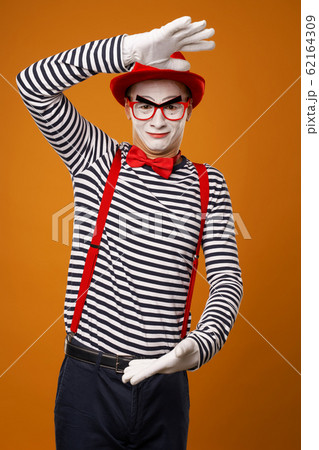 Smiling mime with white face in red hat and striped t-shirt on blank orange background Smiling mime with white face in red hat and striped t-shirt on blank orange background 62164309