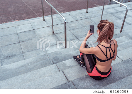 Girl athlete, in summer in city, resting after workout, holding smartphone, reading and writing message on smartphone, listening to music on headphones. Free space for copy text Fitness Motivation. Girl athlete, in summer in city, resting after workout, holding smartphone, reading and writing message on smartphone, listening to music on headphones. Free space for copy text Fitness Motivation. 62166593
