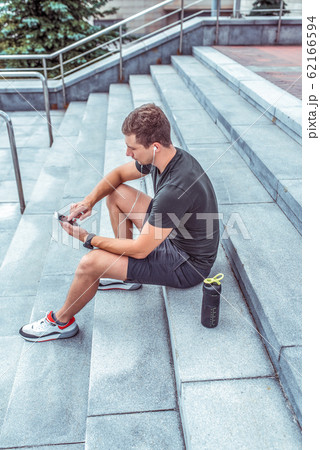male athlete, in summer in city, resting after workout, holding smartphone, reading and writing a message on a smartphone, listening to music on headphones. Bottle shaker with protein and water. male athlete, in summer in city, resting after workout, holding smartphone, reading and writing a message on a smartphone, listening to music on headphones. Bottle shaker with protein and water. 62166594