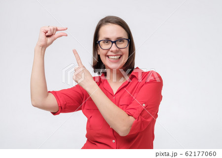 Pretty amazed woman in red shirt and glasses shows something invisible with her fingers. 62177060