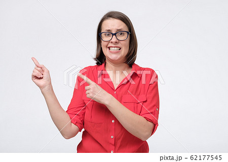 Confused young woman in red shirt and glasses showing aside with finger to copy space place. 62177545
