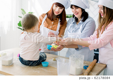 Mothers Day, family, cooking and people concept - little cute baby girl and her mom, aunt and grandmother making and decorating cupcakes at home kitchen Mothers Day, family, cooking and people concept - little cute baby girl and her mom, aunt and grandmother making and decorating cupcakes at home kitchen 62178084