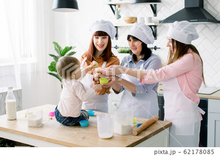 Mothers Day, family, cooking and people concept - little cute baby girl and her mom, aunt and grandmother making and decorating cupcakes at home kitchen Mothers Day, family, cooking and people concept - little cute baby girl and her mom, aunt and grandmother making and decorating cupcakes at home kitchen 62178085