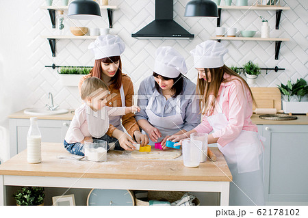 Happy women in white aprons baking together, Cutting out shapes from sugar cookie dough with cookie cutters. Little baby girl helps to make cookies together with mother, aunt and grandmother 62178102