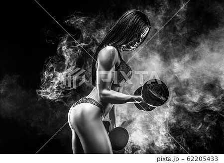 Muscular unrecognizable brunette in bikini doing exercise on biceps with dumbbells in smoke 62205433