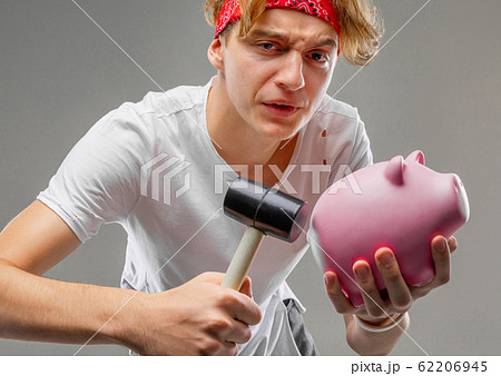 stylish charming blond guy smashing a piggy bank with a hammer on a gray background stylish charming blond guy smashing a piggy bank with a hammer on a gray background 62206945