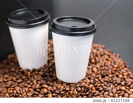 Two White cups of hot coffee on a dark background. Coffee beans background. Vintage style, food banner. Coffee to go in a paper cup, selective focus Two White cups of hot coffee on a dark background. Coffee beans background. Vintage style, food banner. Coffee to go in a paper cup, selective focus 62227106