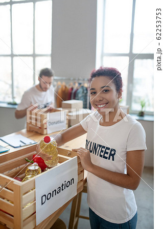 Young volunteer girl standing near food-filled donation box. 62253753