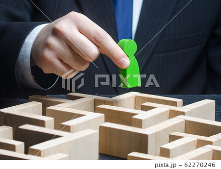 A male businessman holds a green figure over a maze. Make a path to success, provide support. Pass test, complete task. Professional skills development. Find a way out of the situation. Reach the goal 62270246