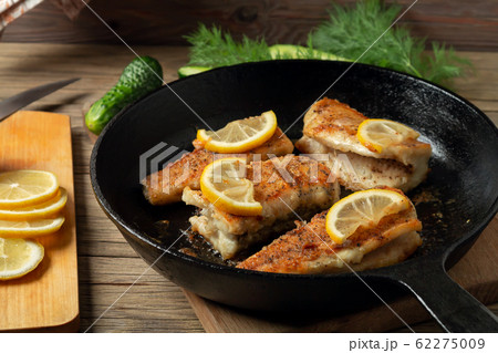Portioned pieces of fried fish in a black cast-iron pan with fresh cucumbers and herbs. Traditional dinner of a village fisherman Portioned pieces of fried fish in a black cast-iron pan with fresh cucumbers and herbs. Traditional dinner of a village fisherman 62275009