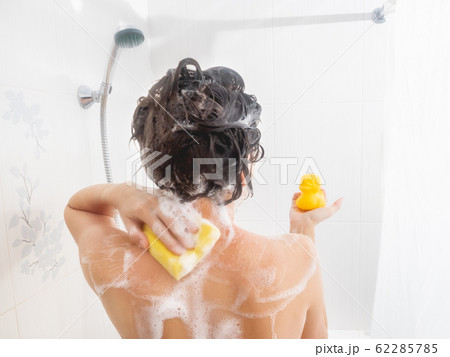 Naked woman with short hair takes a shower with funny yellow rubber duck. Woman washes her shoulders with yellow sponge. White bathroom. Naked woman with short hair takes a shower with funny yellow rubber duck. Woman washes her shoulders with yellow sponge. White bathroom. 62285785