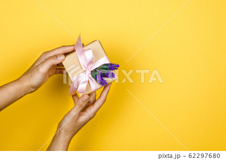 close-up woman hands holding a gift box with a lavender twig in a gesture of giving on yellow background with copy space 62297680