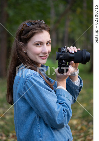 Cute hipster photographer girl takes a picture on a professional camera and smiles. Portrait in the autumn park, against the background of trees and fallen leaves Cute hipster photographer girl takes a picture on a professional camera and smiles. Portrait in the autumn park, against the background of trees and fallen leaves 62300715