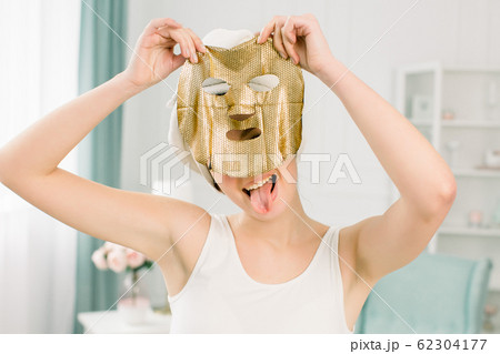 Cosmetic procedure, pretty funny girl in white towel with gold sheet mask on light background. Beauty, spa, skin and body care. Cosmetic procedure, pretty funny girl in white towel with gold sheet mask on light background. Beauty, spa, skin and body care. 62304177