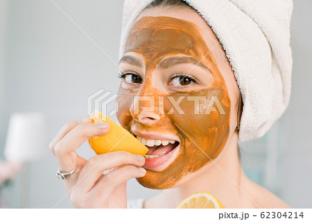 Attractive young woman with white towel on the head and with mud facial brown mask bites a lemon and smiles. Beauty, spa, skin and body care. Attractive young woman with white towel on the head and with mud facial brown mask bites a lemon and smiles. Beauty, spa, skin and body care. 62304214