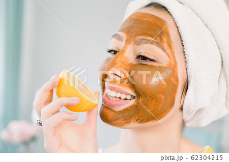 Attractive young woman with white towel on the head and with mud facial brown mask bites a lemon and smiles. Beauty, spa, skin and body care. Attractive young woman with white towel on the head and with mud facial brown mask bites a lemon and smiles. Beauty, spa, skin and body care. 62304215