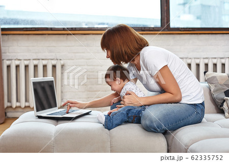 A woman with a child at a laptop sitting on a sofa. Work at home, freelancer, work during maternity leave for remote access. 62335752
