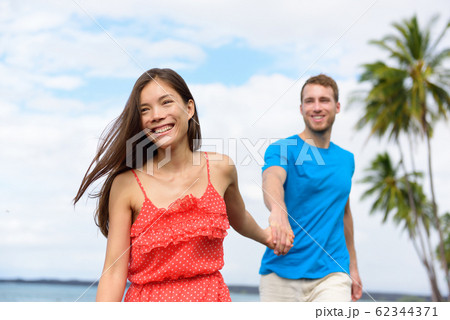Happy summer vacation couple walking holding hands on beach travel destination. Asian woman caucasian man multiracial people together having fun 62344371