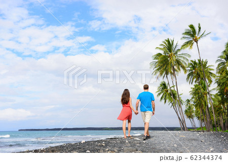 Couple walking on Hawaii secluded black sand volcanic beach on the Big island of Hawaii, hawaiian destination tropical travel for summer vacations. People from behind relaxing on walk Couple walking on Hawaii secluded black sand volcanic beach on the Big island of Hawaii, hawaiian destination tropical travel for summer vacations. People from behind relaxing on walk 62344374