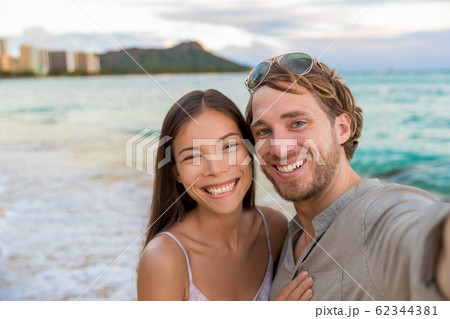 Selfie couple on Waikiki beach taking pictures with smartphone during night out walk on beach summer vacations in Honolulu, Hawaii. Travel destination. Young people having fun on hawaiian holidays 62344381
