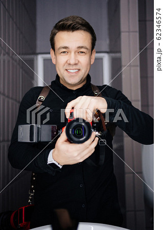 Photographer in black clothes taking a selfie with the camera in the mirror and smiling Photographer in black clothes taking a selfie with the camera in the mirror and smiling 62346574