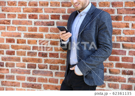 Young casual business man using smartphone in urban city background relaxing on brick wall texting sms on phone app living a modern lifestyle. Closeup of unrecognizable person 62361135