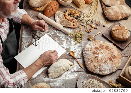 Old Baker writing down old-time recipe in bakery notebook surrounded by bread 62382769