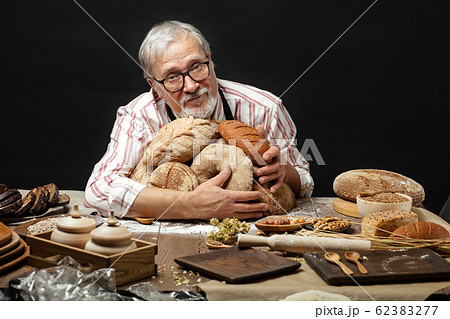 Happy old baker looking at camera and smiling while hugging loaves of bread 62383277