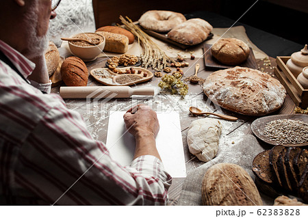 Old Baker writing down old-time recipe in bakery notebook surrounded by bread Old Baker writing down old-time recipe in bakery notebook surrounded by bread 62383828