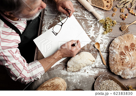 Old Baker writing down old-time recipe in bakery notebook surrounded by bread Old Baker writing down old-time recipe in bakery notebook surrounded by bread 62383830
