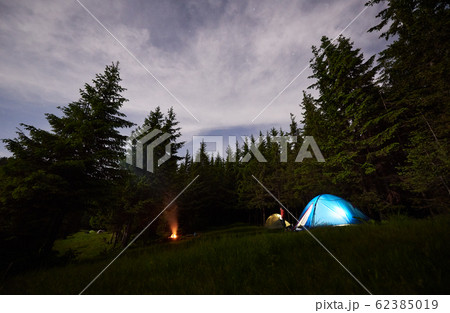 Camping in forest at the evening. Tents and burning bonfire among green fir trees under starry sky with haze of smoke. Camping in forest at the evening. Tents and burning bonfire among green fir trees under starry sky with haze of smoke. 62385019
