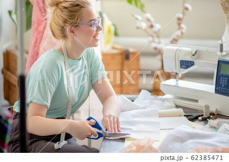 Beautiful focused young fashion designer cutting pink lace fabric in workshop 62385471