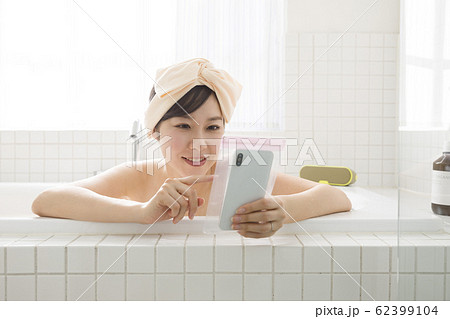 バスルームで防水ケースにスマホを入れて操作する若い女性 バスルームで防水ケースにスマホを入れて操作する若い女性 62399104