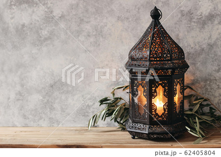 Black ornamental Moroccan, Arabic lantern. Green olive leaves, branches on old wooden table, blurred grunge wall background,. Greeting card for Muslim holiday Ramadan Kareem. Festive still life. 62405804