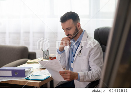 Dark-haired bearded doctor in a white robe looking concentrated Dark-haired bearded doctor in a white robe looking concentrated 62411611