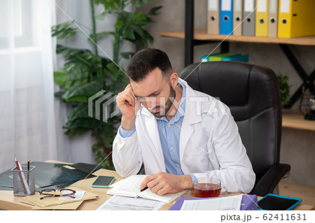 Young bearded doctor in a white robe looking involved reading the report Young bearded doctor in a white robe looking involved reading the report 62411631