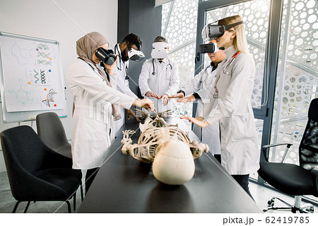 Education, science, vr, medical and health concept. Group of multiethnic doctors, biologists, archeologists, scientists wearing VR goggles study human skeleton in modern laboratory 62419785