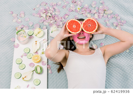 Young cheerful european woman with pink mud facial mask lies on bed and holds big grapefruits, covering her eyes. Healthy diet, skincare, facial treatment, cosmetology concept. 62419936