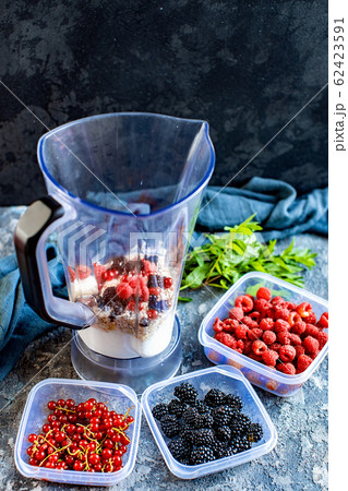 Blender with oatmeal, plastic plates, containers with raspberry, blackberry, red currant, cherry on a stone background. Making a healthy smoothie on a blender 62423591
