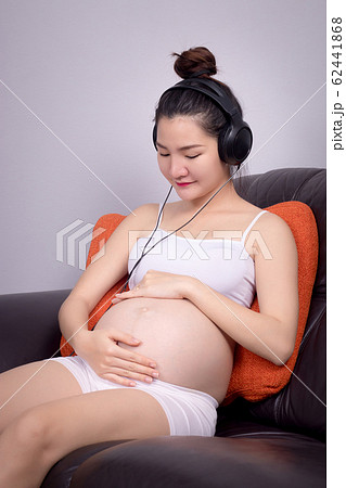 Portrait of pregnant Asian woman sitting on sofa at home and listening music in headphones. Beautiful Asia female model in her 20s. 62441868