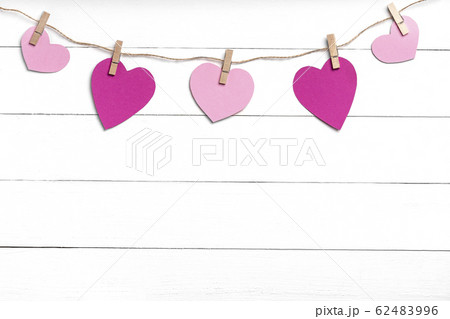 Clothespins with pink hearts hanging on rope behind white wooden surface. Clothespins with pink hearts hanging on rope behind white wooden surface. 62483996